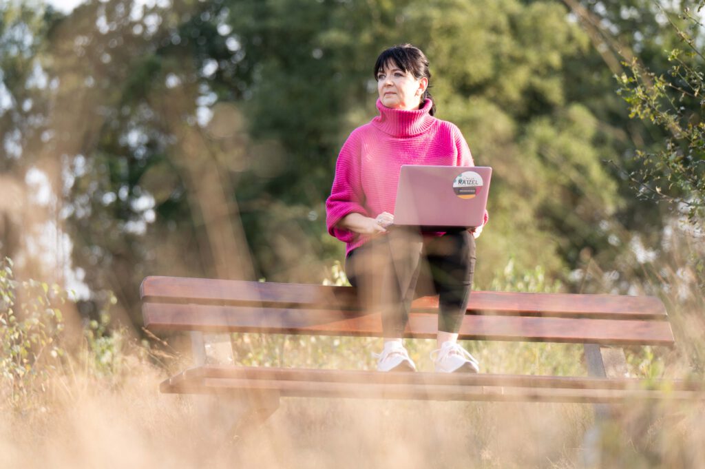 Frau in pinkem Pulli mit Laptop auf Bank in der Natur sitzend